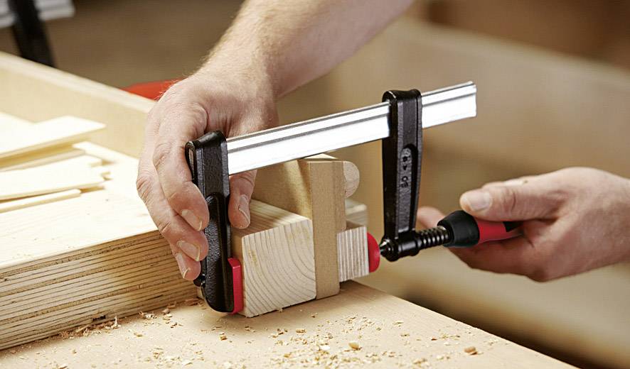 Una persona blocca un pezzo di legno su un banco da lavoro utilizzando una morsa per segare o levigare. Trucioli di legno sono visibili.