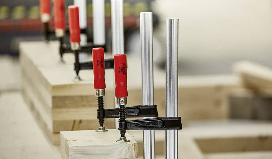 Diverse morse con impugnature rosse tengono insieme travi di legno su un banco da lavoro, pronte per l'assemblaggio o l'incollaggio.