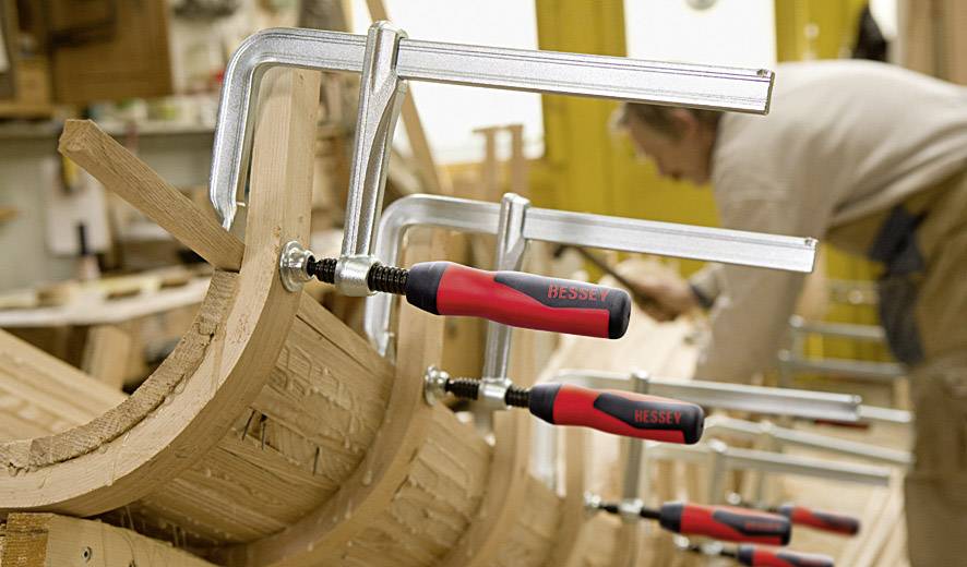 Un falegname lavora in un laboratorio e fissa pezzi di legno con diverse morse. Il focus è sulle morse, sullo sfondo sfocato.