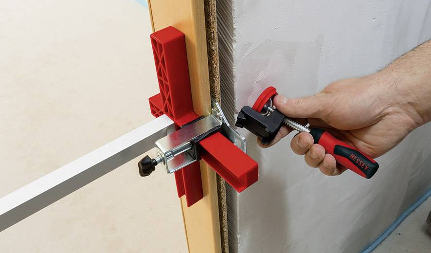 Una mano regola una morsa rossa su un telaio della porta utilizzando una chiave inglese. Una striscia metallica stabilizza la struttura.