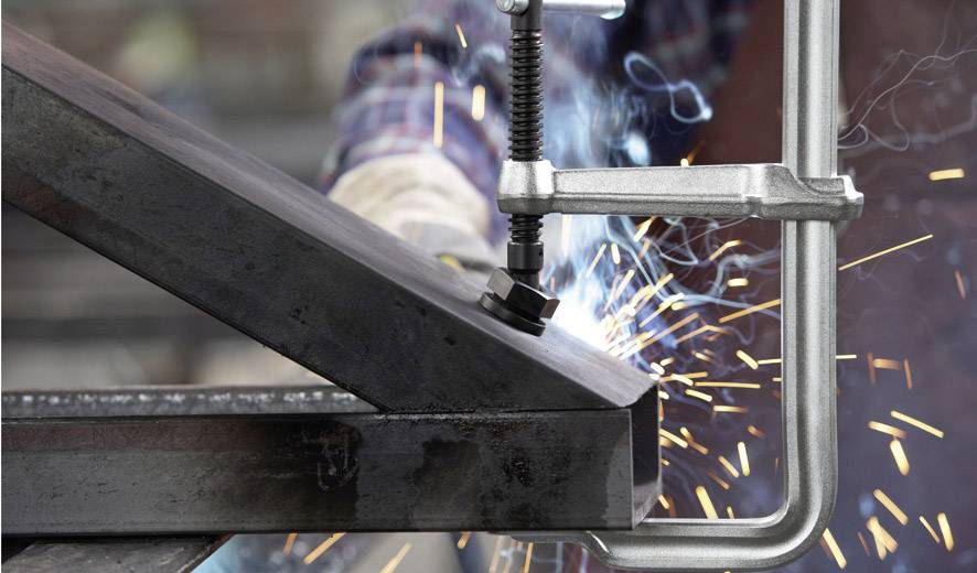 Una persona in abbigliamento da lavoro salda due pezzi di metallo ad angolo retto. Scintille volano mentre una morsa tiene fermi i pezzi.