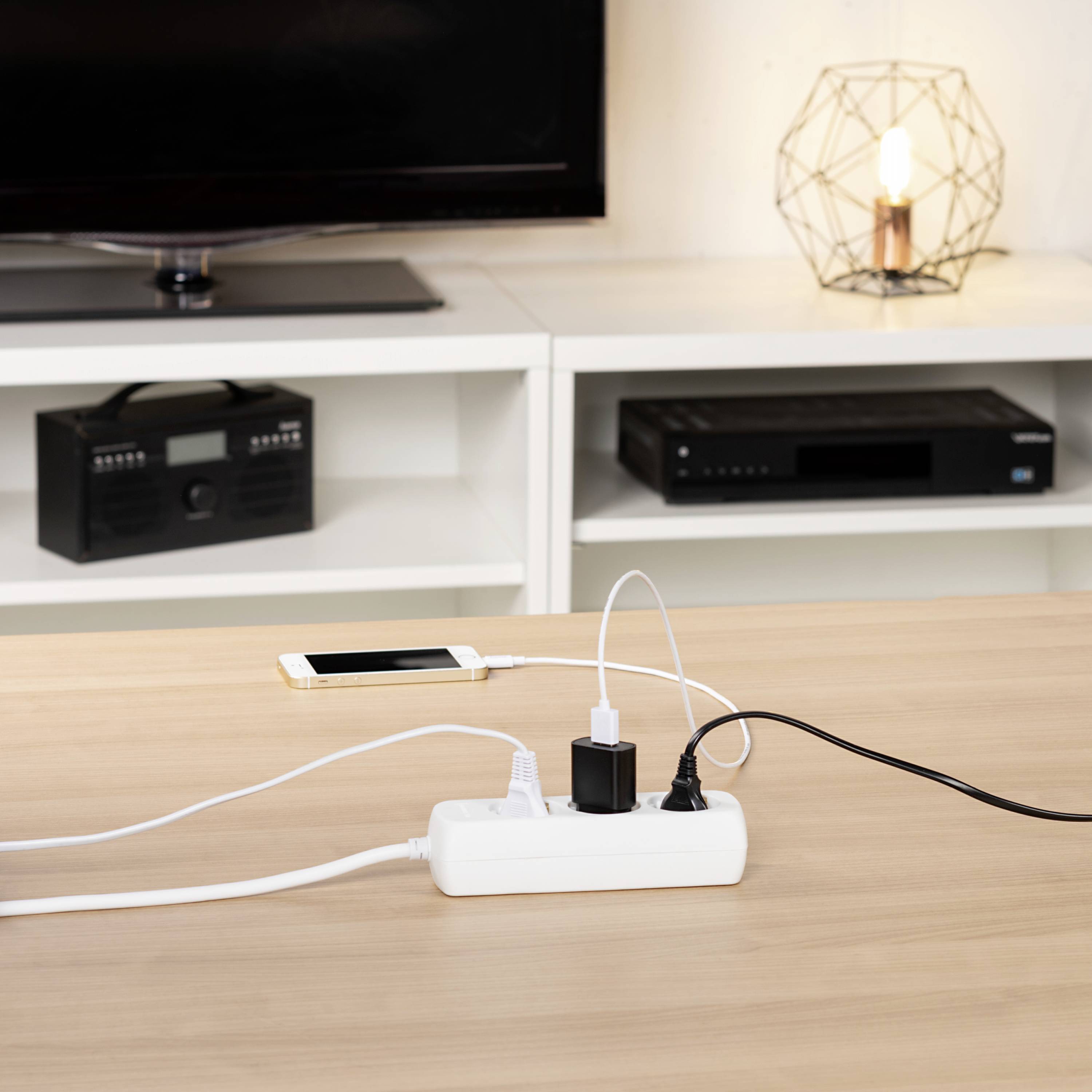 Un tavolo con una presa multipla in cui sono inseriti diversi dispositivi come smartphone e lampada. Sullo sfondo un televisore e una radio.