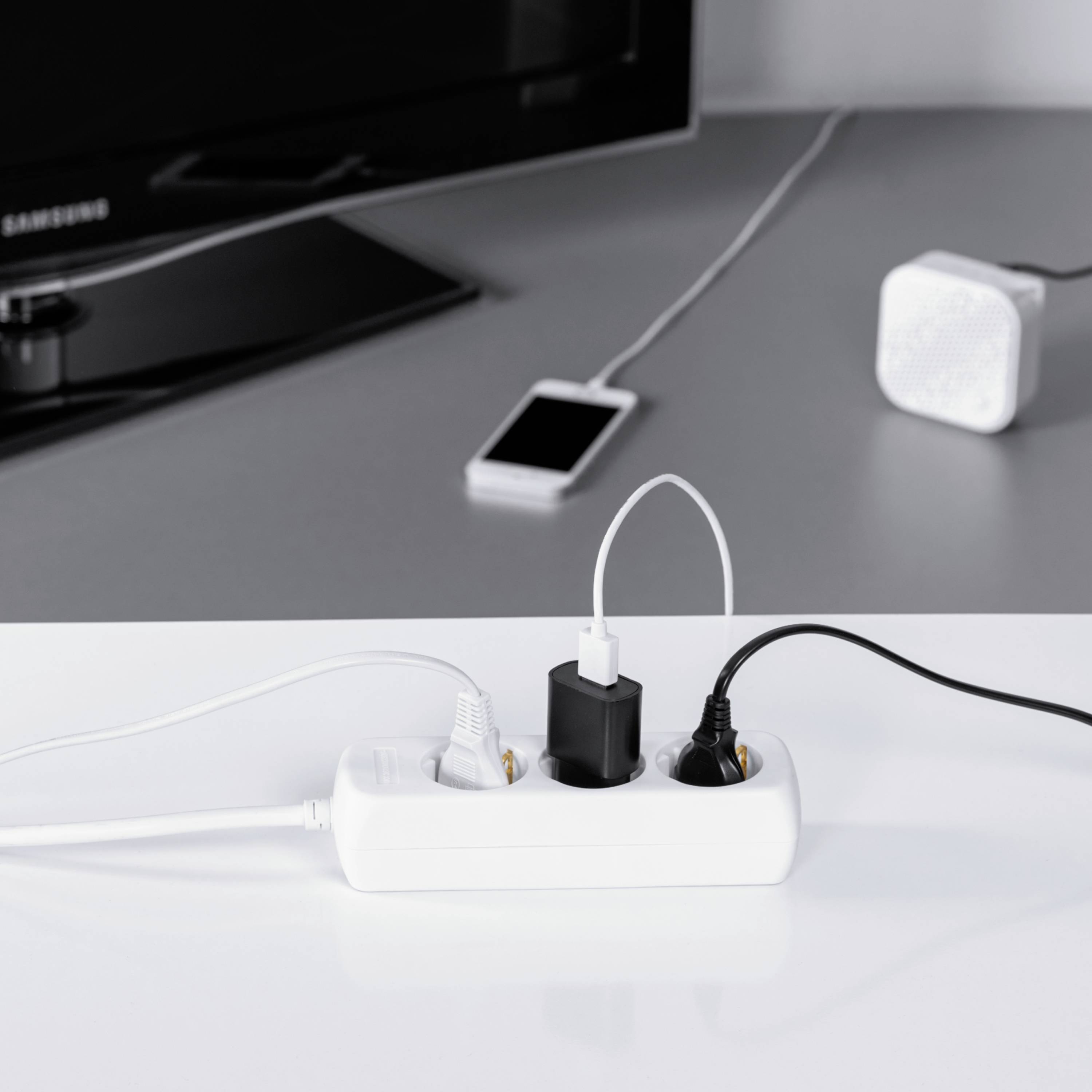 Multipresa con tre caricabatterie inseriti su un tavolo, accanto uno smartphone e un altoparlante sullo sfondo.
