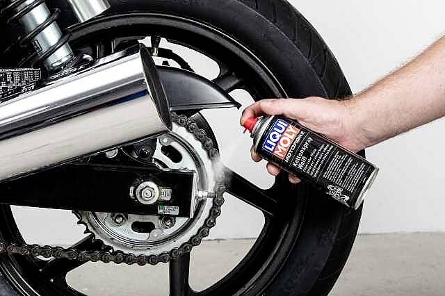 Una mano spruzza detergente per catene da un barattolo spray sulla catena di una moto nera.