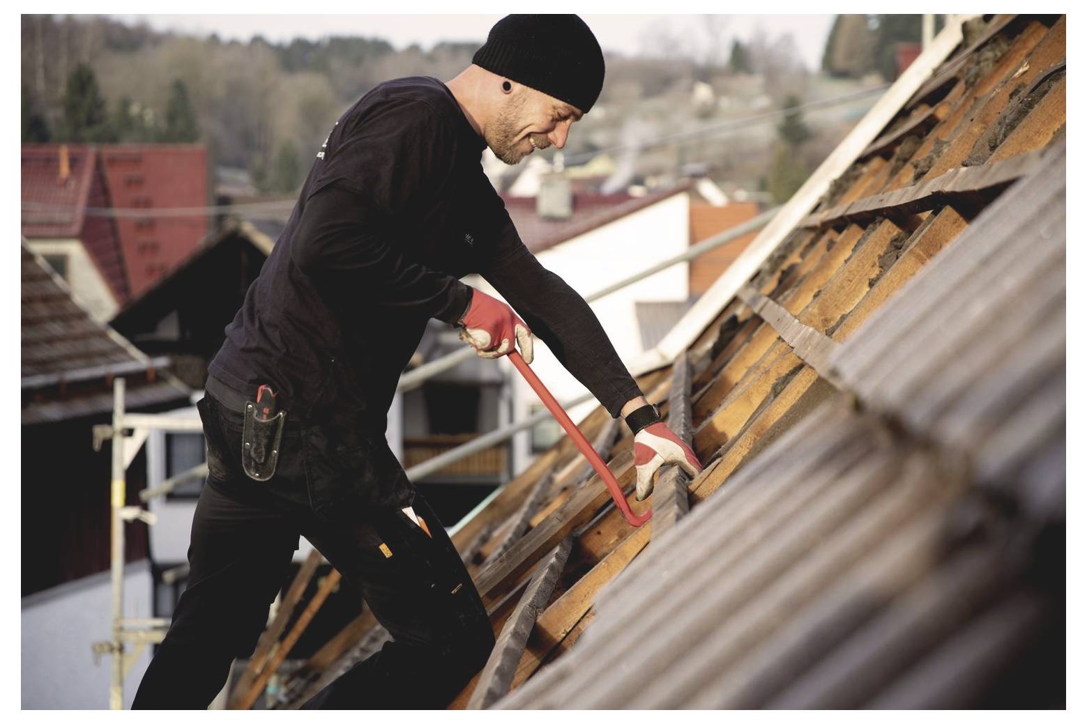 Un uomo con un completo nero e un berretto utilizza una palanchina per riparare un tetto inclinato, circondato da altri tetti e alberi sullo sfondo.