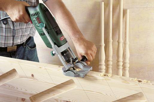 Una persona sta utilizzando una fresatrice elettrica per tagliare con precisione degli intagli in una trave di legno. Sullo sfondo ci sono quattro aste tornite in legno.