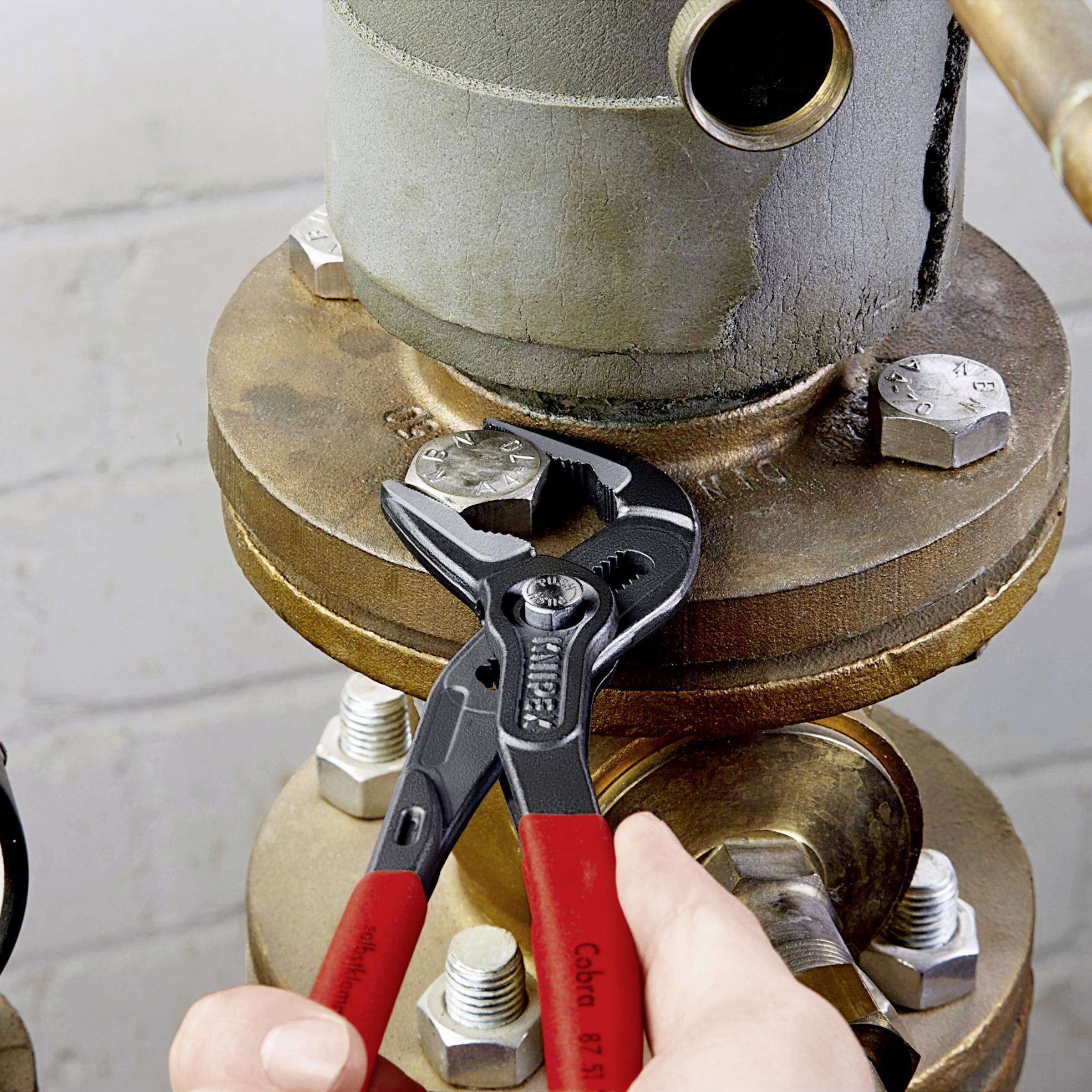 Una mano tiene una pinza rossa e svita un bullone su un grande connettore metallico con tubo. Sullo sfondo: un muro di mattoni grigi.