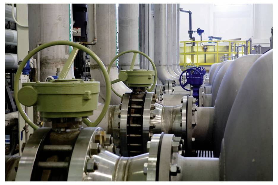 Tubi e valvole di controllo in un impianto industriale, che mostrano ruote delle valvole verdi e blu e grandi tubi metallici sullo sfondo luminoso.