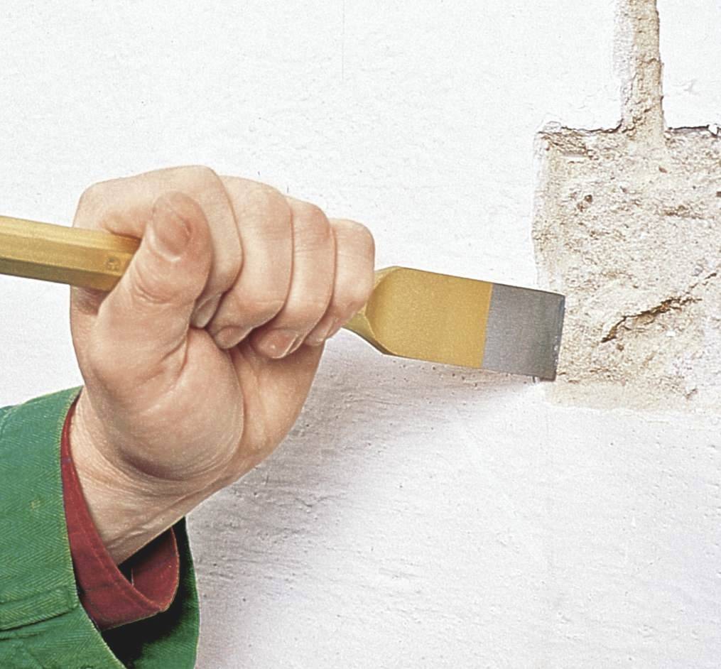 Una mano tiene una spatola e rimuove l'intonaco da una parete bianca. Una parte della parete è già scoperta.