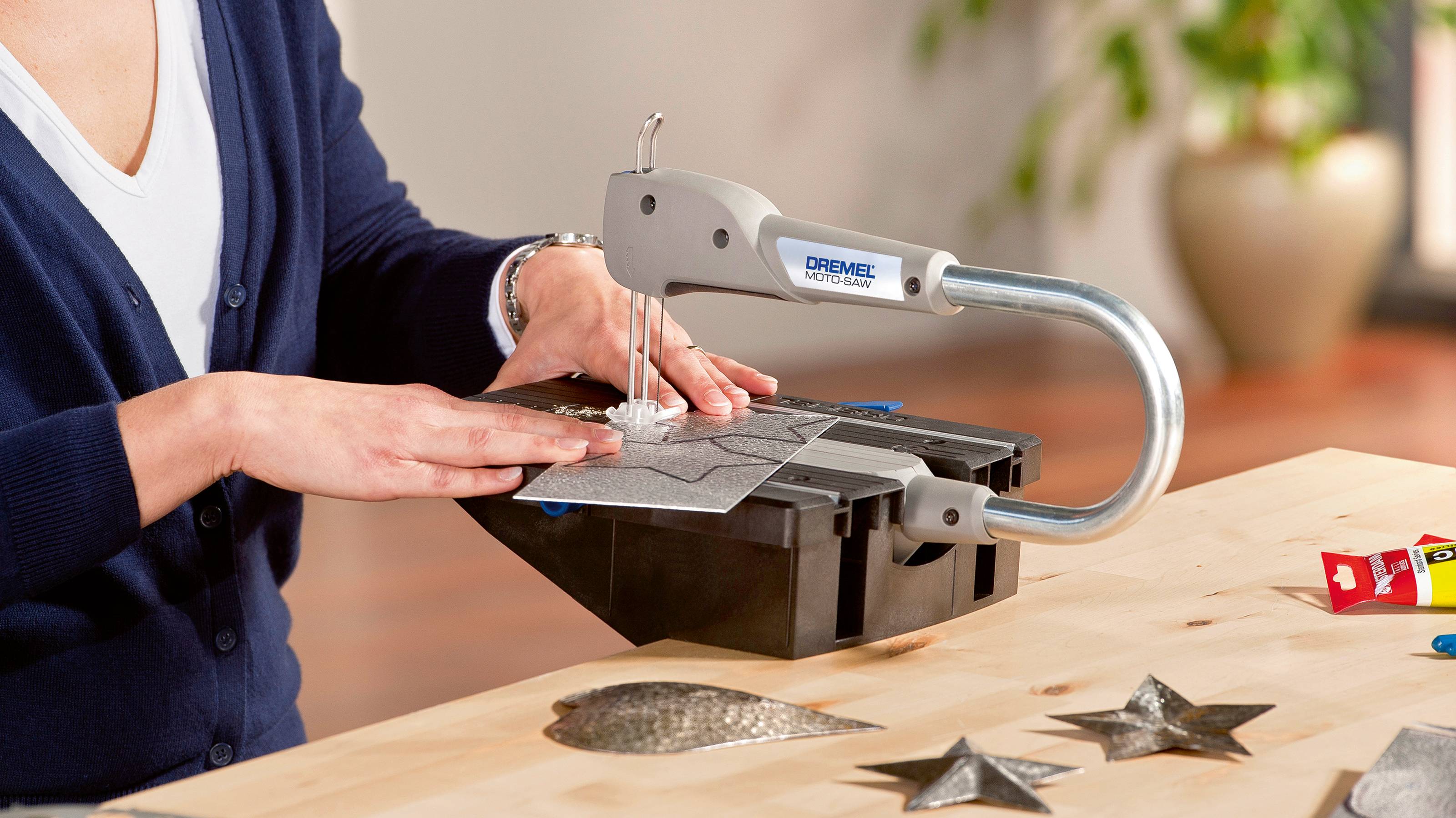 Una persona sta utilizzando un Dremel Moto-Saw per tagliare una lastra grigia. Sul tavolo ci sono forme metalliche già ritagliate.