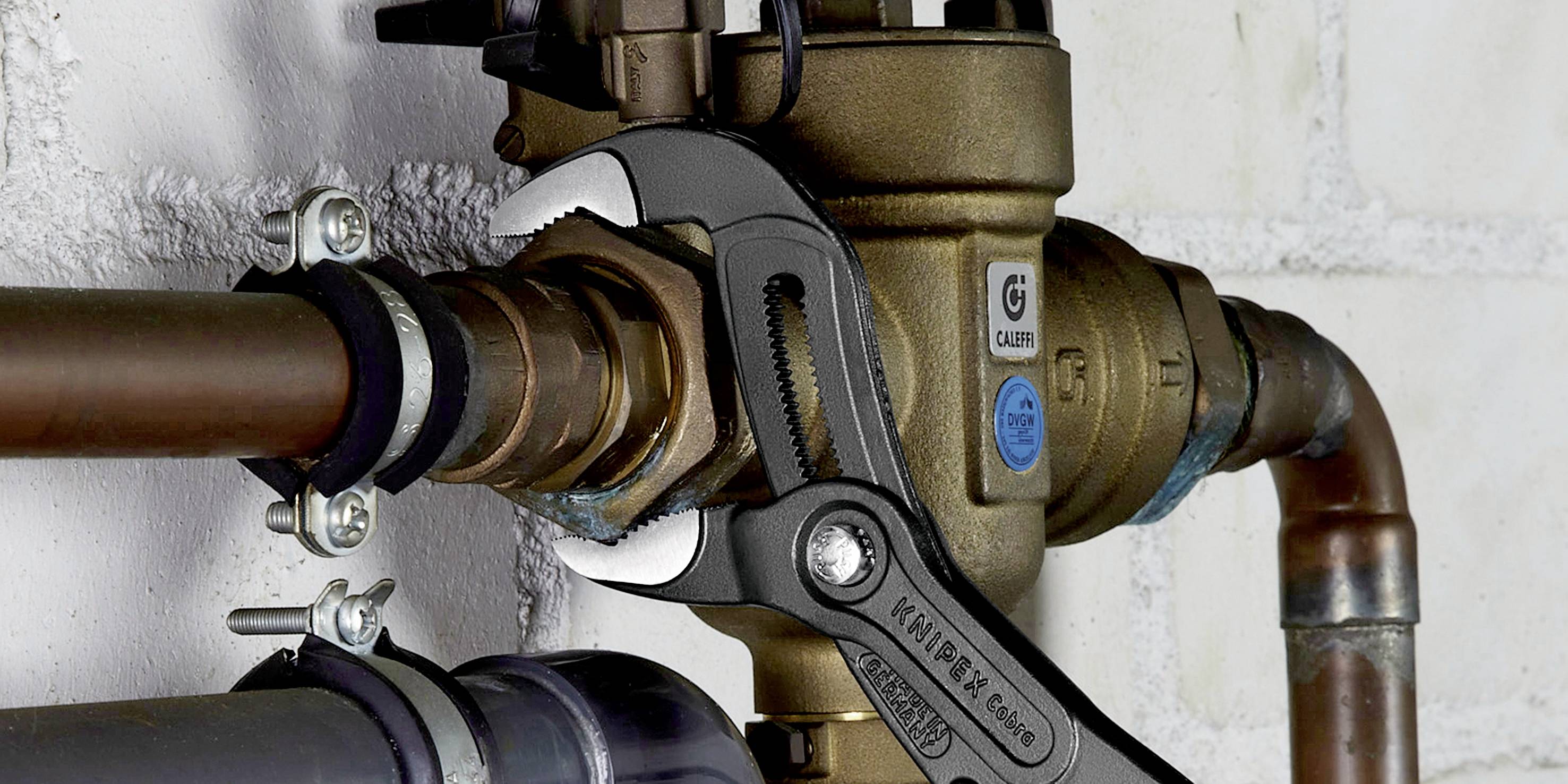 Una chiave regolabile afferra saldamente un giunto metallico in un sistema di tubazioni fissato a una parete di mattoni bianchi.