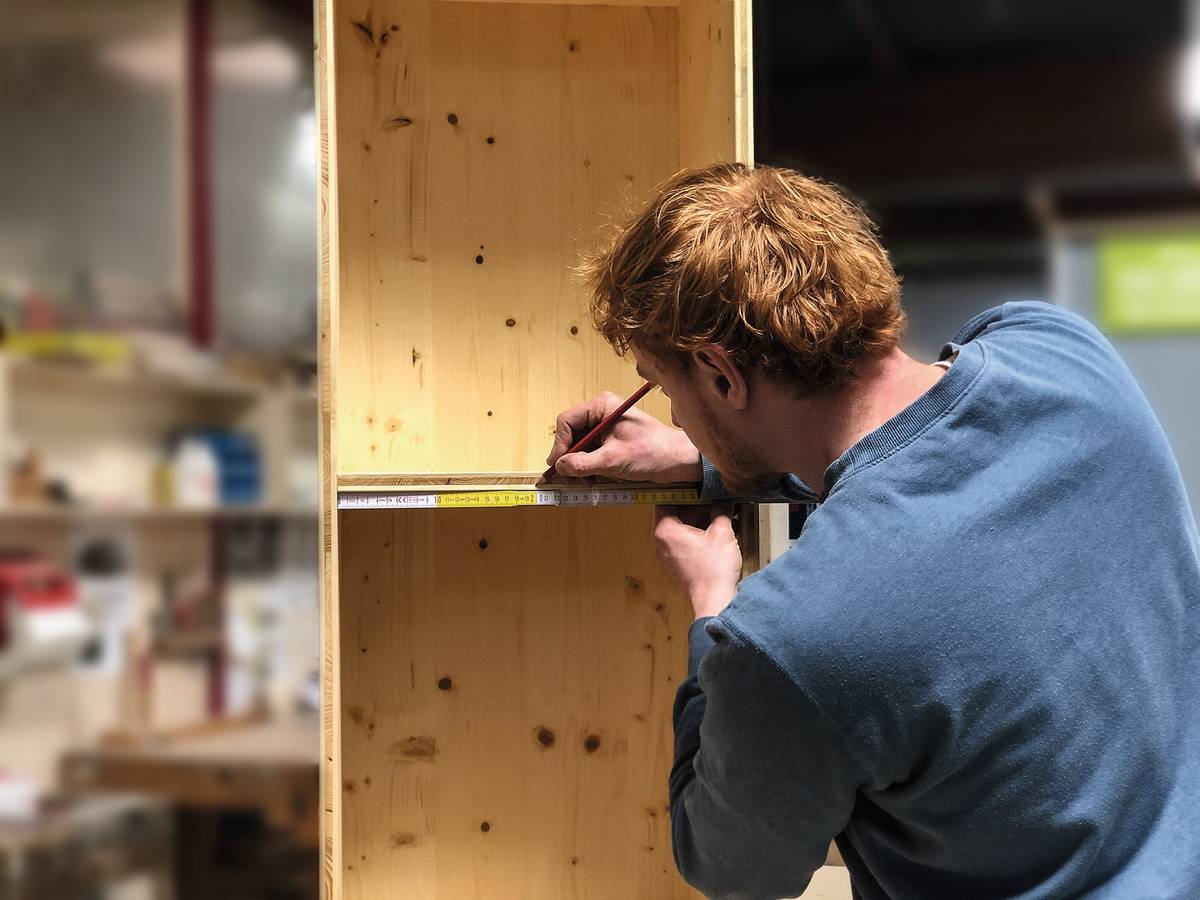 Una persona misura con un nastro metrico e segna il legno di uno scaffale in un ambiente di officina.