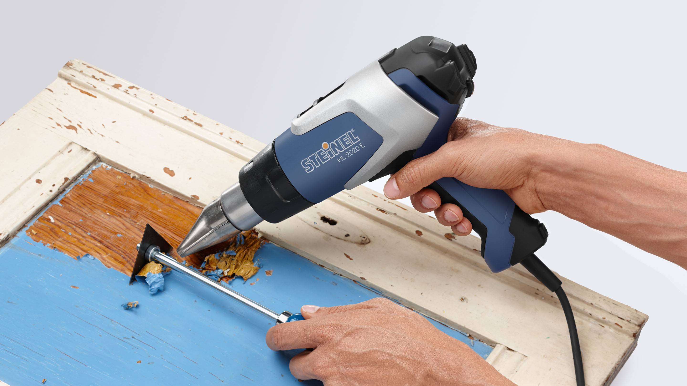 Una persona usa una pistola termica blu per rimuovere la vernice da un vecchio telaio di una porta in legno.