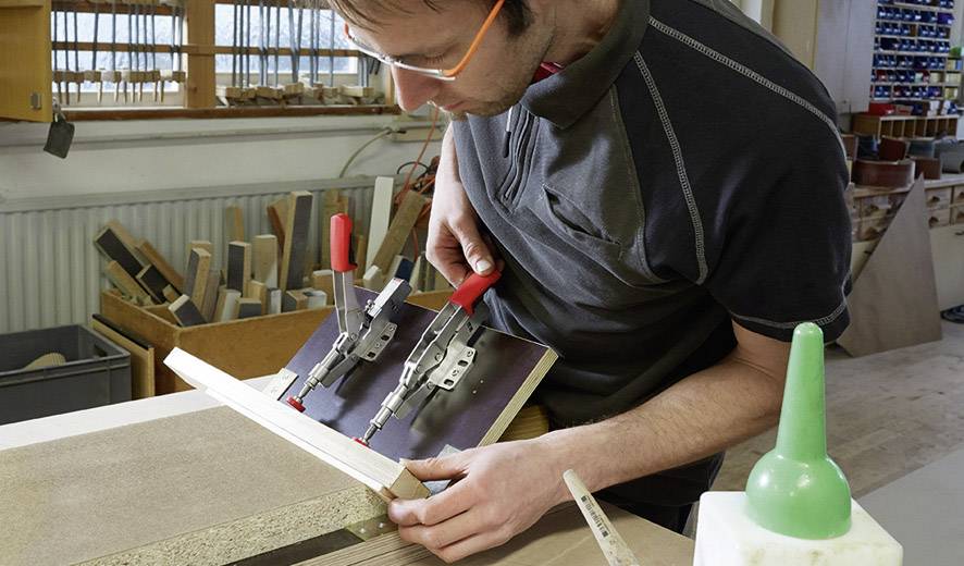 Un uomo lavora in un laboratorio e fissa dei pezzi di legno con una morsa. Sullo sfondo sono visibili scaffali con altri materiali.