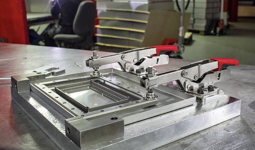 Lavorazione dei metalli: Un telaio metallico stabile con due morsetti rossi su un tavolo da lavoro in un ambiente di officina.