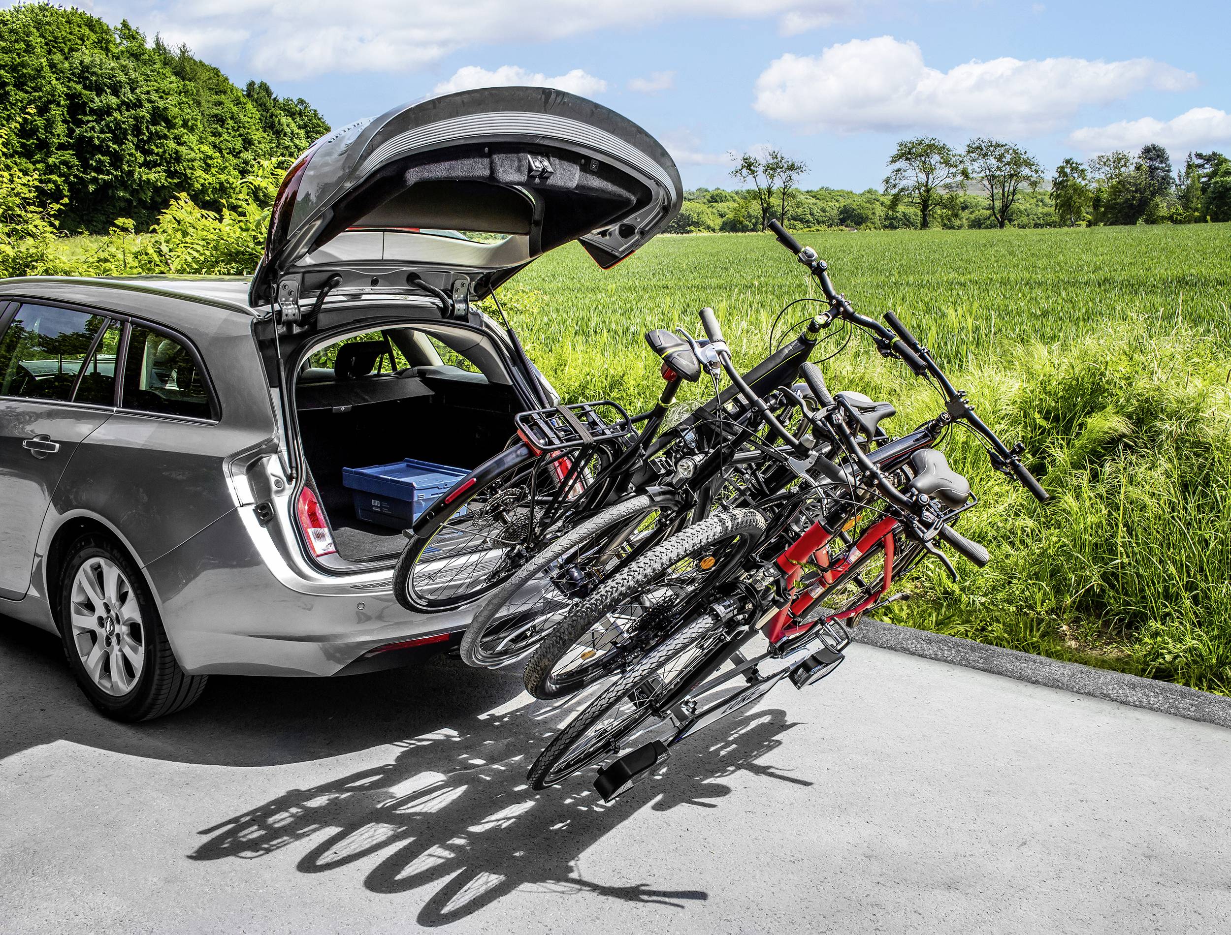 Un station wagon è parcheggiato su un sentiero di campagna con il portellone posteriore aperto, un portabiciclette con quattro biciclette fissato, sullo sfondo campi verdi.