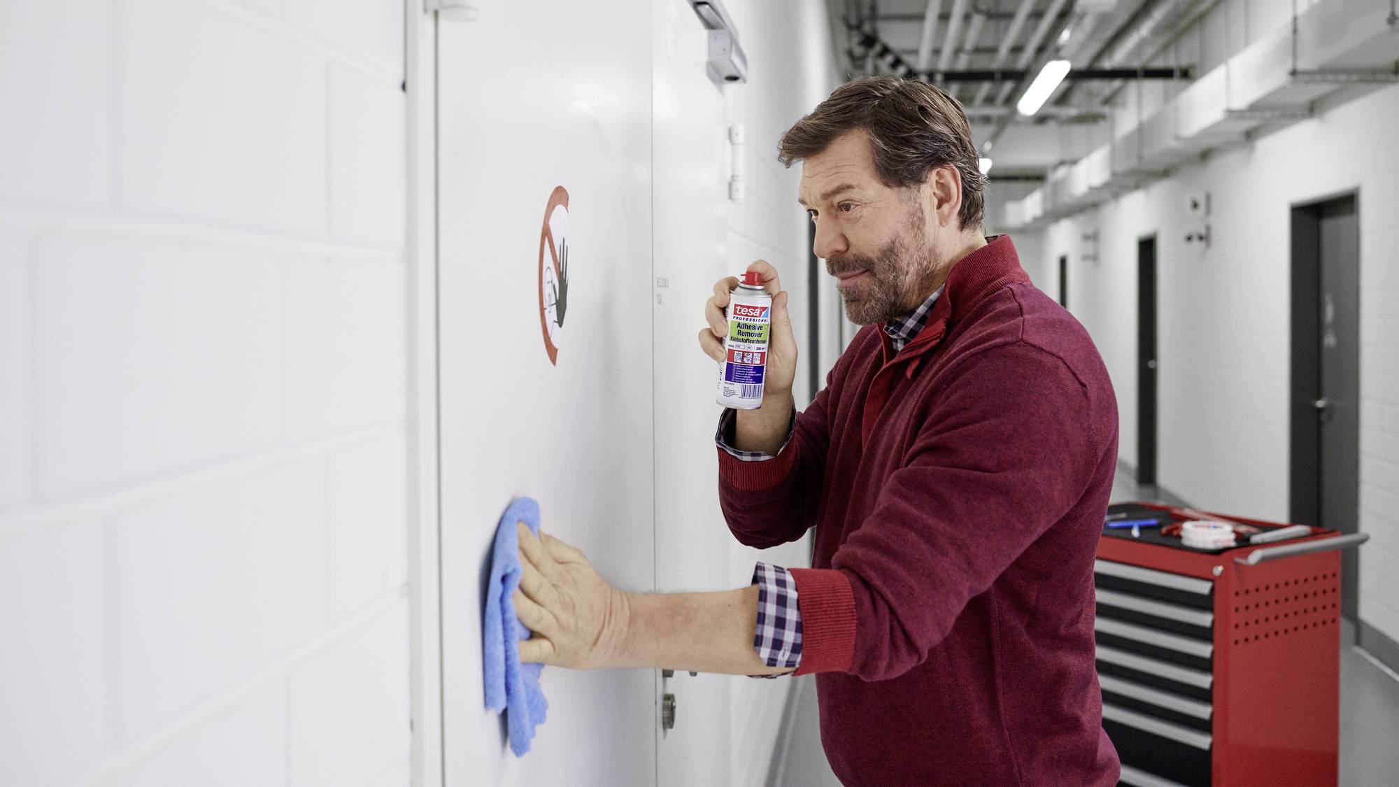 Un uomo sta pulendo accuratamente una parete metallica con un detergente e un panno in un corridoio industriale. Sullo sfondo è visibile un carrello degli attrezzi.