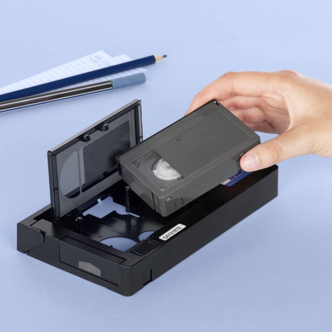 Una mano inserisce una cassetta VHS in un videoregistratore nero. Sullo sfondo ci sono un blocco note, una penna a sfera e una matita.