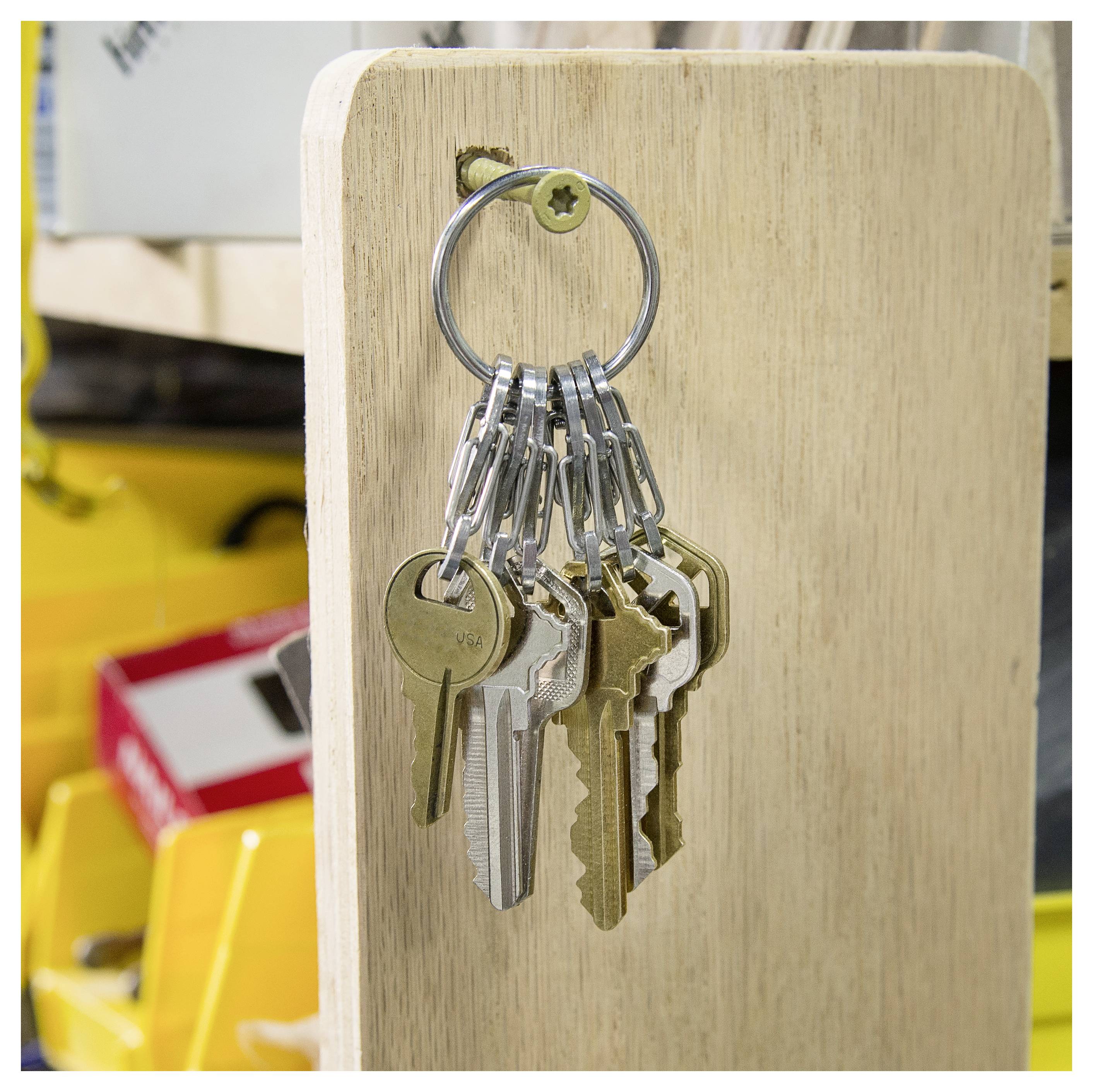 Un insieme di varie chiavi è appeso a un anello metallico fissato a una tavola di legno, con uno sfondo sfocato di attrezzi e scatole.