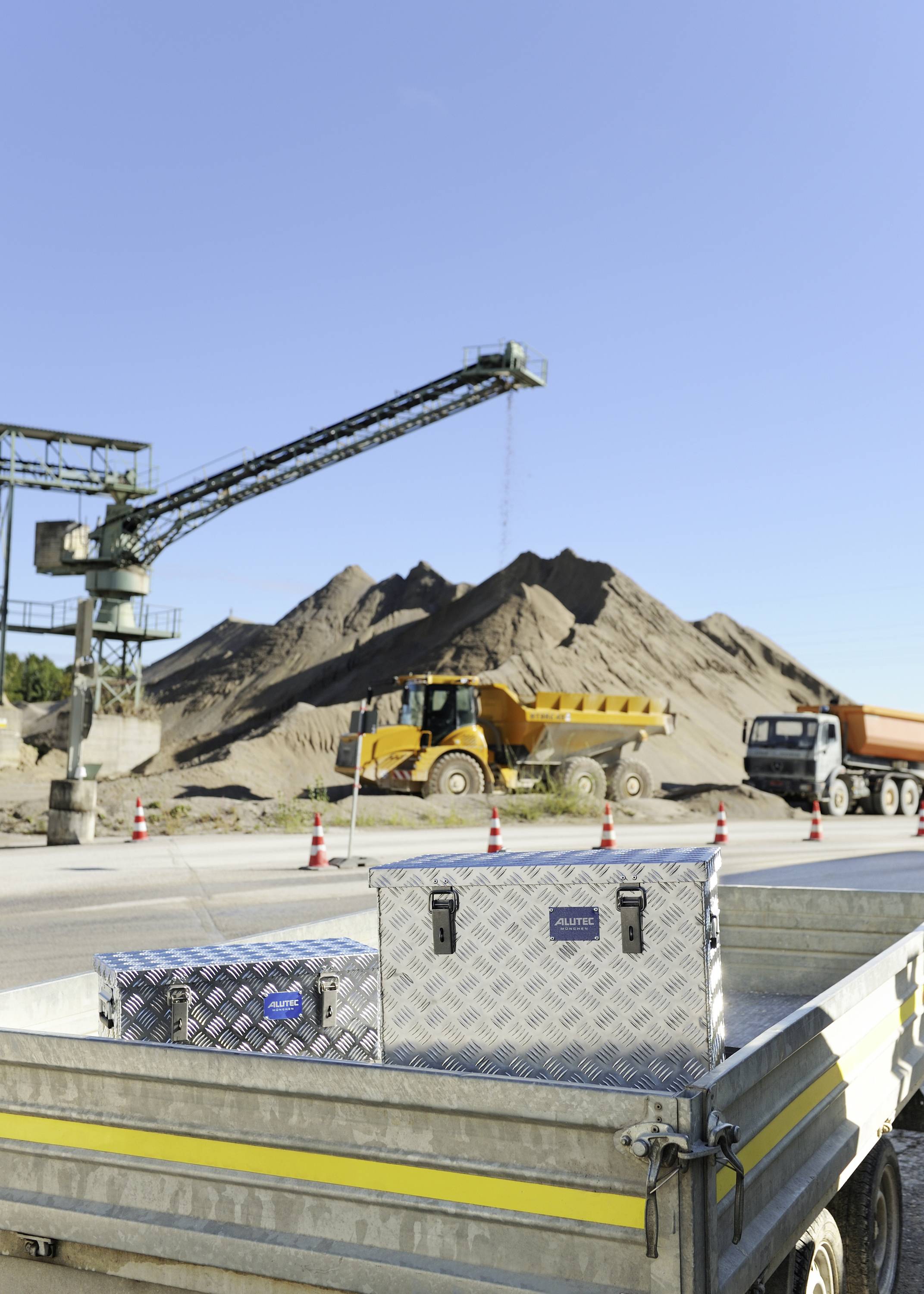 Un grande cumulo di sabbia in un cantiere edile con un camion e un mezzo da lavoro sullo sfondo. In primo piano, un rimorchio aperto carico di scatole.