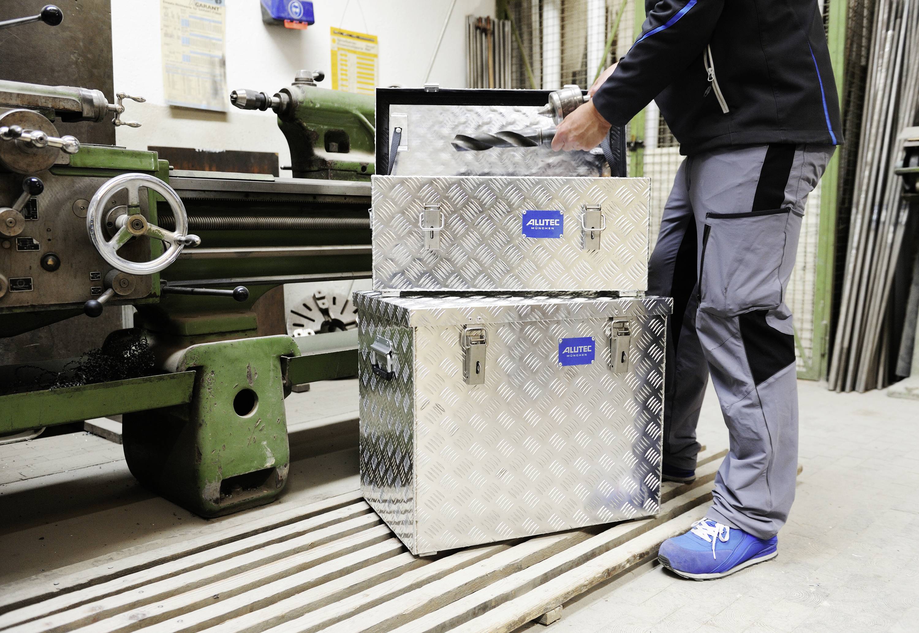 Una persona con abiti da lavoro sta impilando scatole metalliche argentate in un'officina accanto a una grande macchina verde.