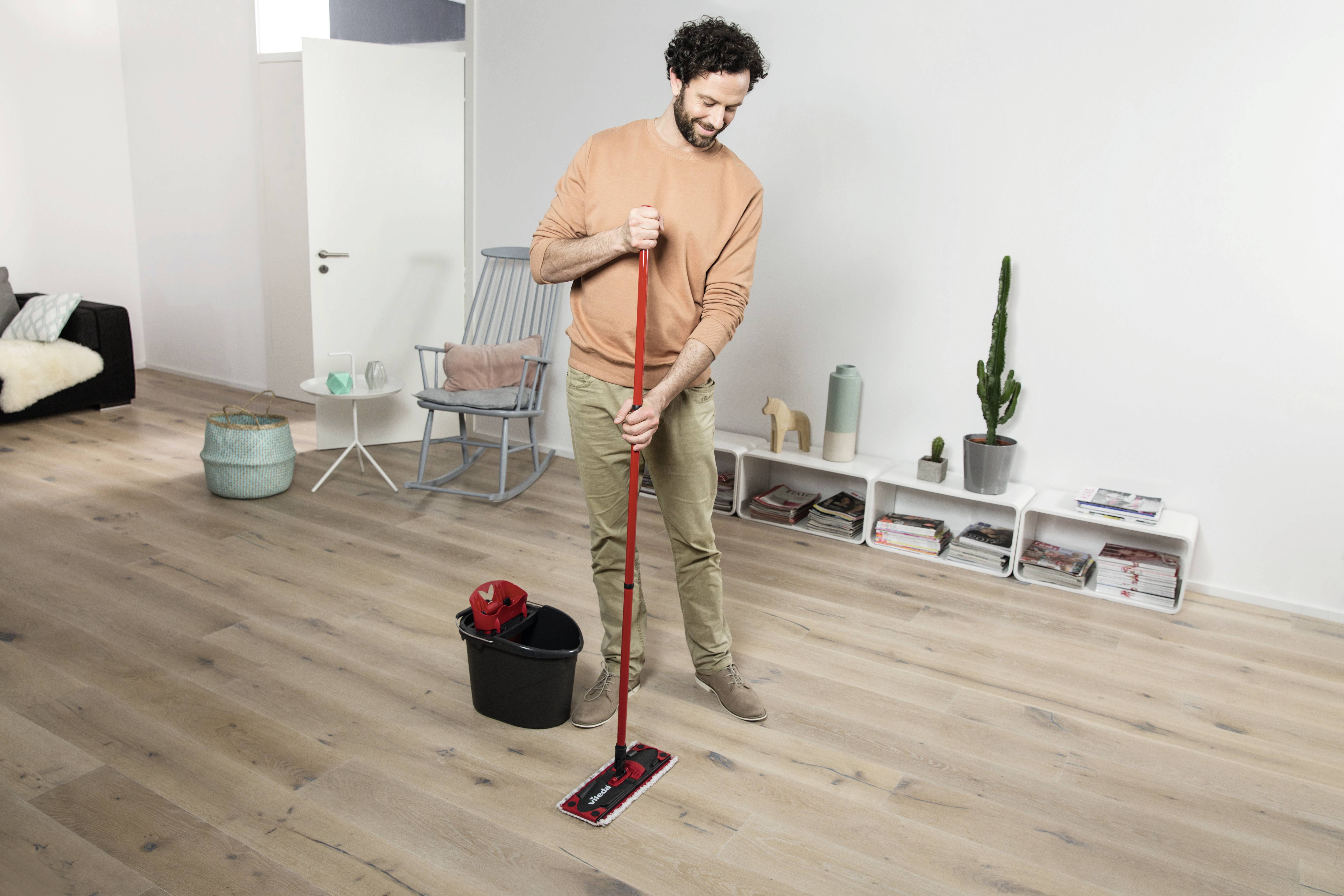 Un uomo sta pulendo il pavimento in legno di un soggiorno moderno con un mocio rosso; sullo sfondo sono visibili un secchio e alcuni mobili.