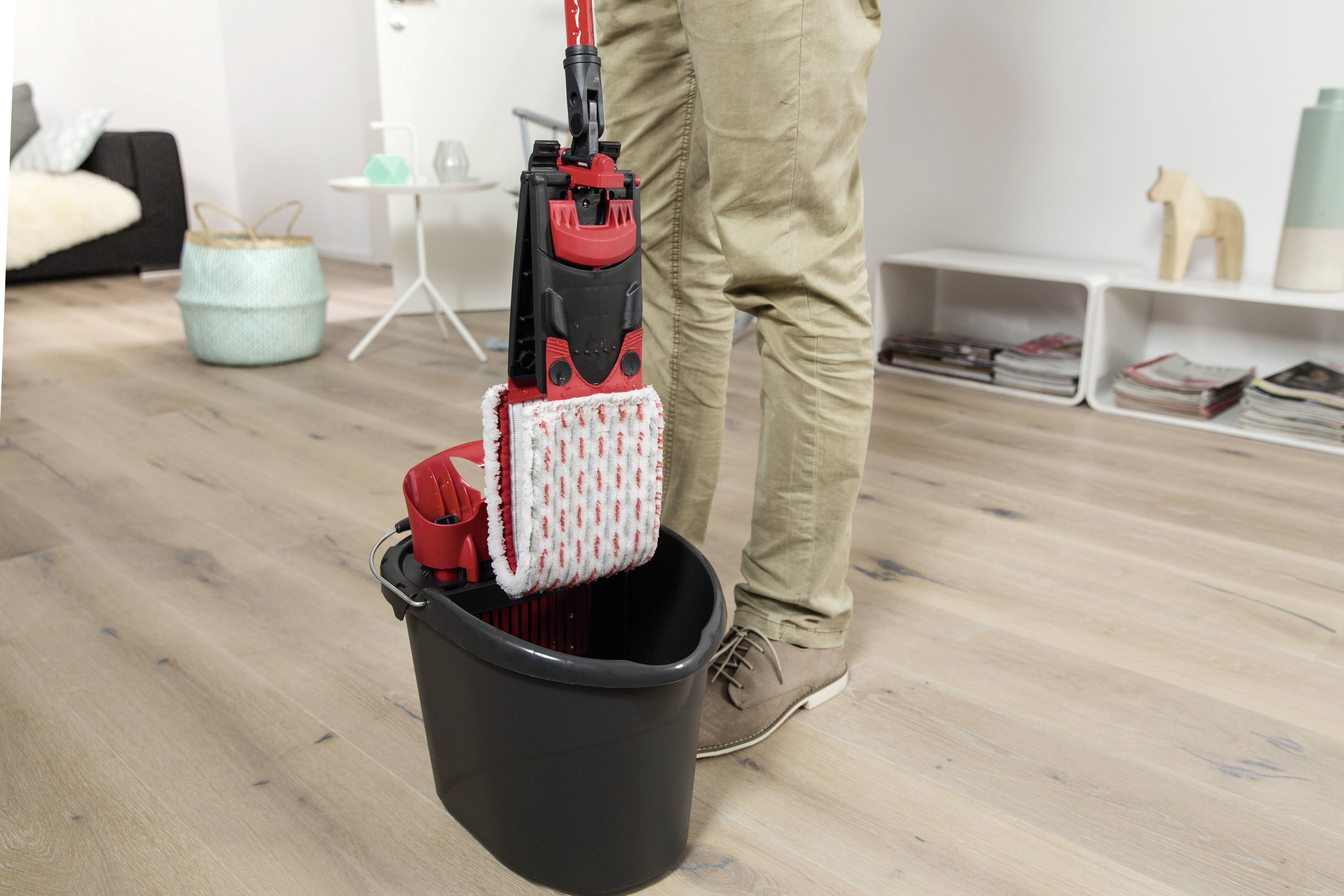 Una persona sta pulendo il pavimento con un mocio e un secchio in un soggiorno. Sullo sfondo sono visibili un divano e una libreria.