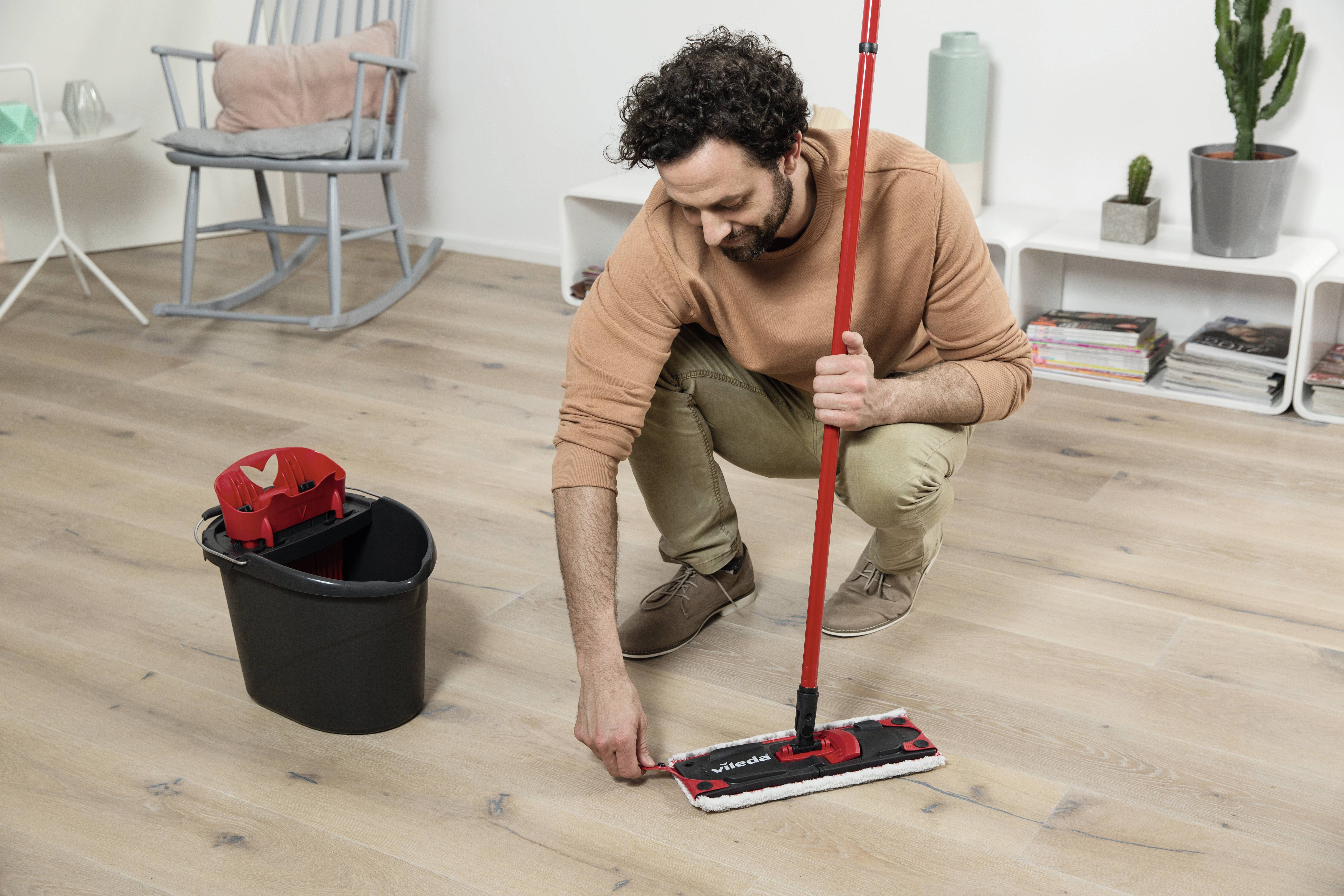 Un uomo sta pulendo un pavimento in legno con un mocio. Un secchio è posizionato accanto a lui. Sullo sfondo sono visibili una poltrona e una scaffalatura.