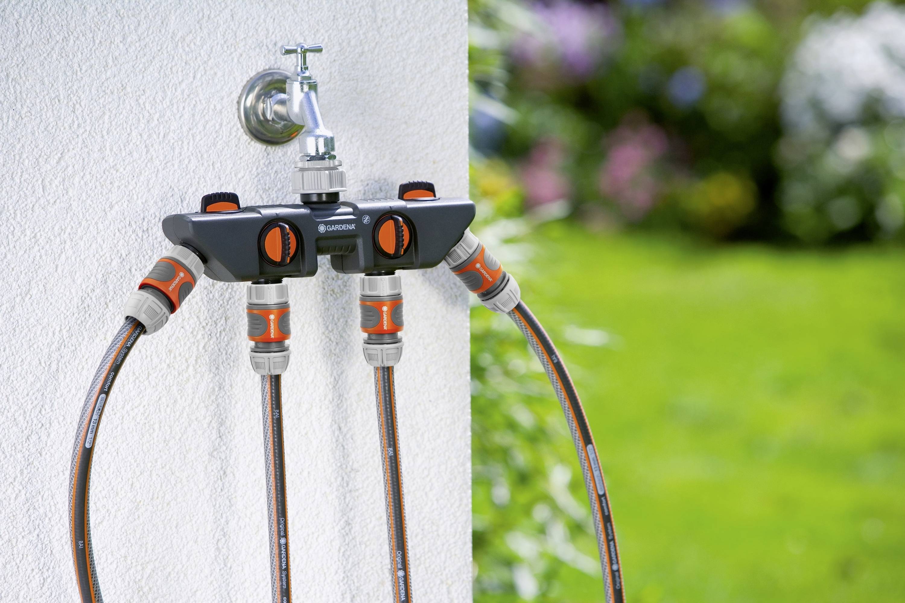 Distributore per tubo da giardino con quattro attacchi montato su una parete esterna, collegato a tubi flessibili, circondato da prato verde e fiori colorati.