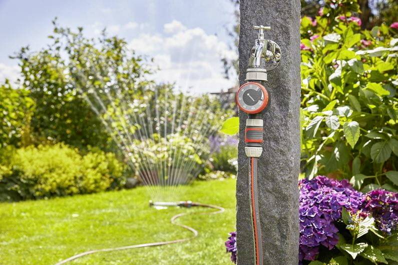 Un timer per l'irrigazione su un rubinetto regola automaticamente l'irrigazione del giardino. Sullo sfondo, uno spruzzatore per il prato spruzza acqua.