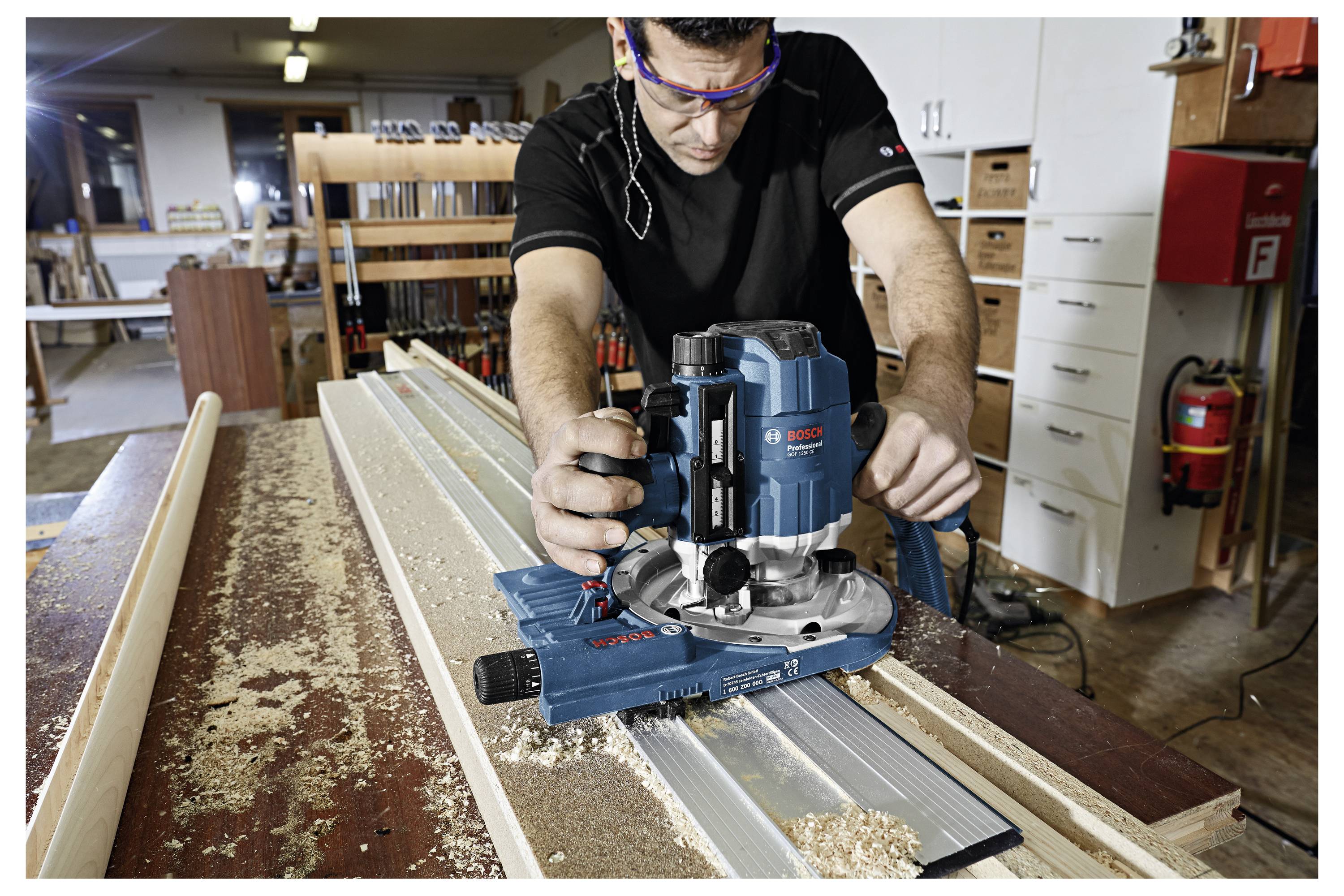 Artigiano utilizza un utensile elettrico per fresare in un laboratorio di falegnameria, lavorando una lista in legno. Trucioli di sega sul tavolo, scaffali sullo sfondo.