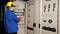 Un elettricista in tuta da lavoro blu e casco giallo sta controllando i quadri elettrici con un multimetro in una sala di controllo.