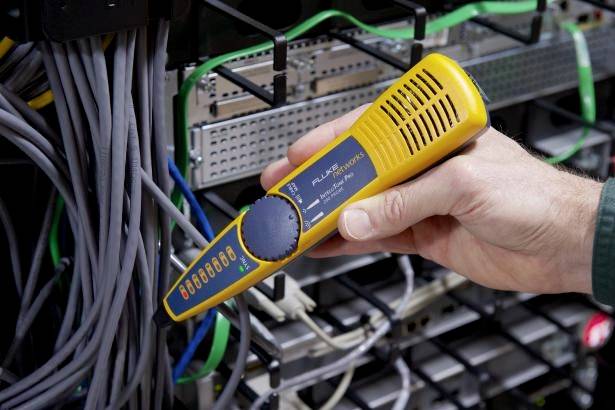 Una persona tiene un dispositivo di test di rete giallo in una sala server, circondato da cavi e hardware. Il dispositivo mostra l'indicazione di funzionamento.