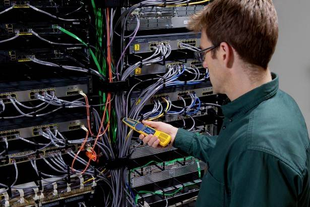 Un tecnico in abbigliamento verde sta controllando i connettori dei cavi con uno strumento di misurazione in una sala server.