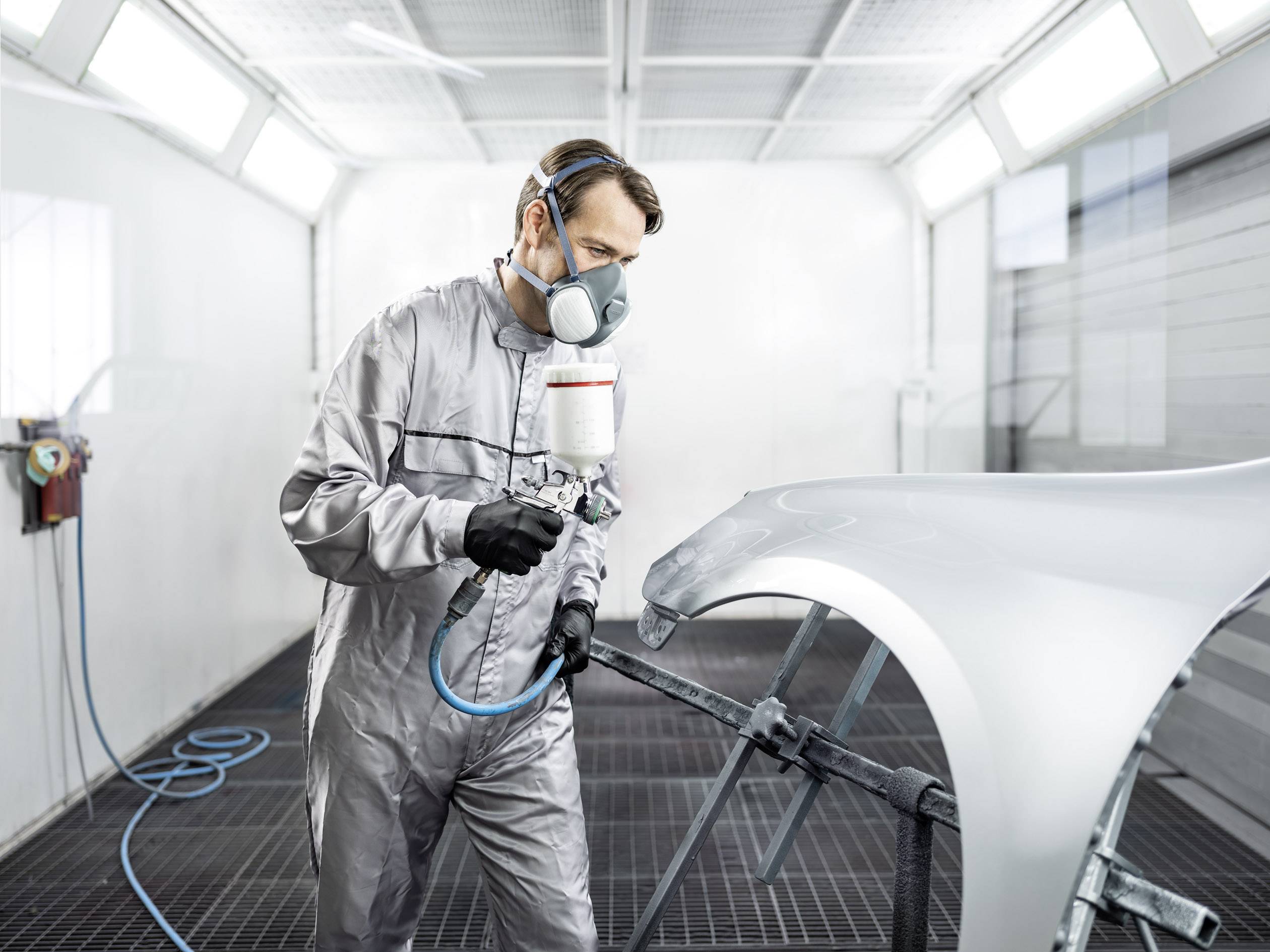 Un uomo in tuta protettiva e maschera respiratoria vernicia un componente auto in una cabina di verniciatura professionale.