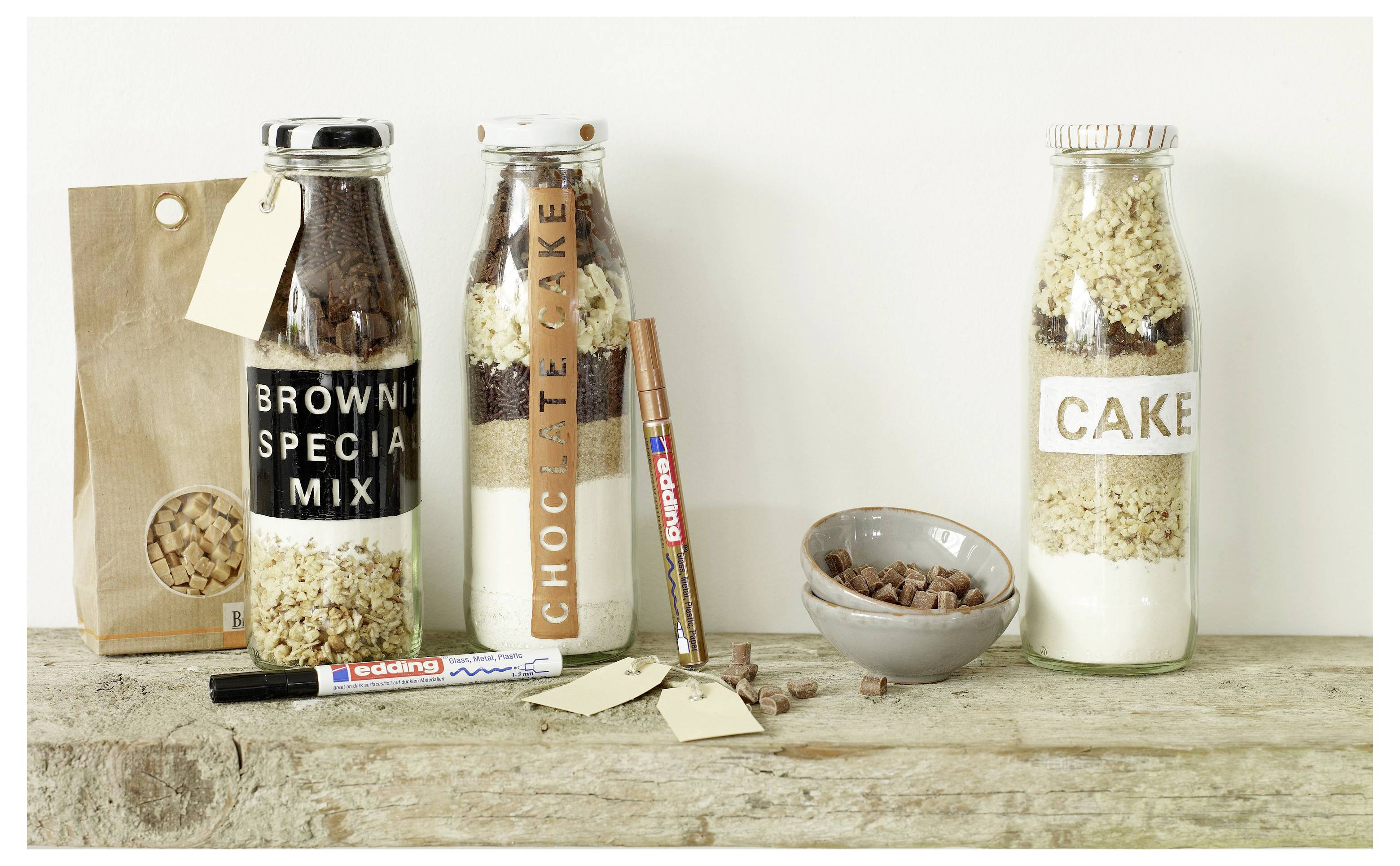 Tre barattoli regalo con miscele di dolci su un tavolo: miscela per brownie, miscela per torta al cioccolato e miscela per torta, con penne, etichette e una borsa di biscotti.