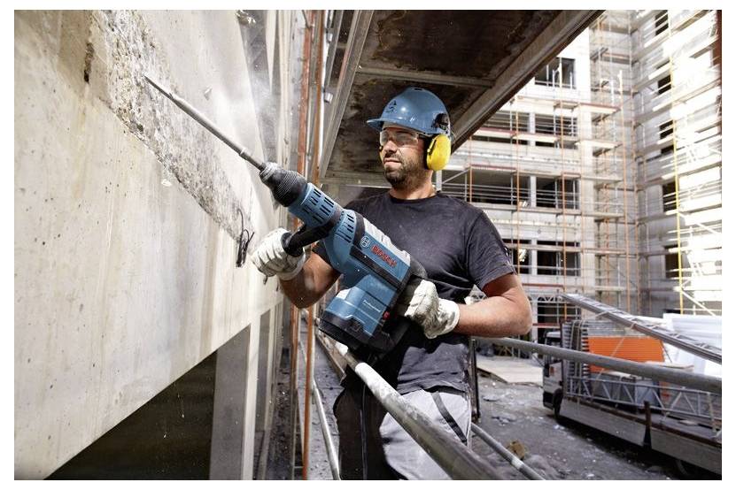 Un lavoratore edile con dispositivi di sicurezza utilizza un trapano elettrico su una parete in calcestruzzo in un cantiere, circondato da impalcature e materiali.