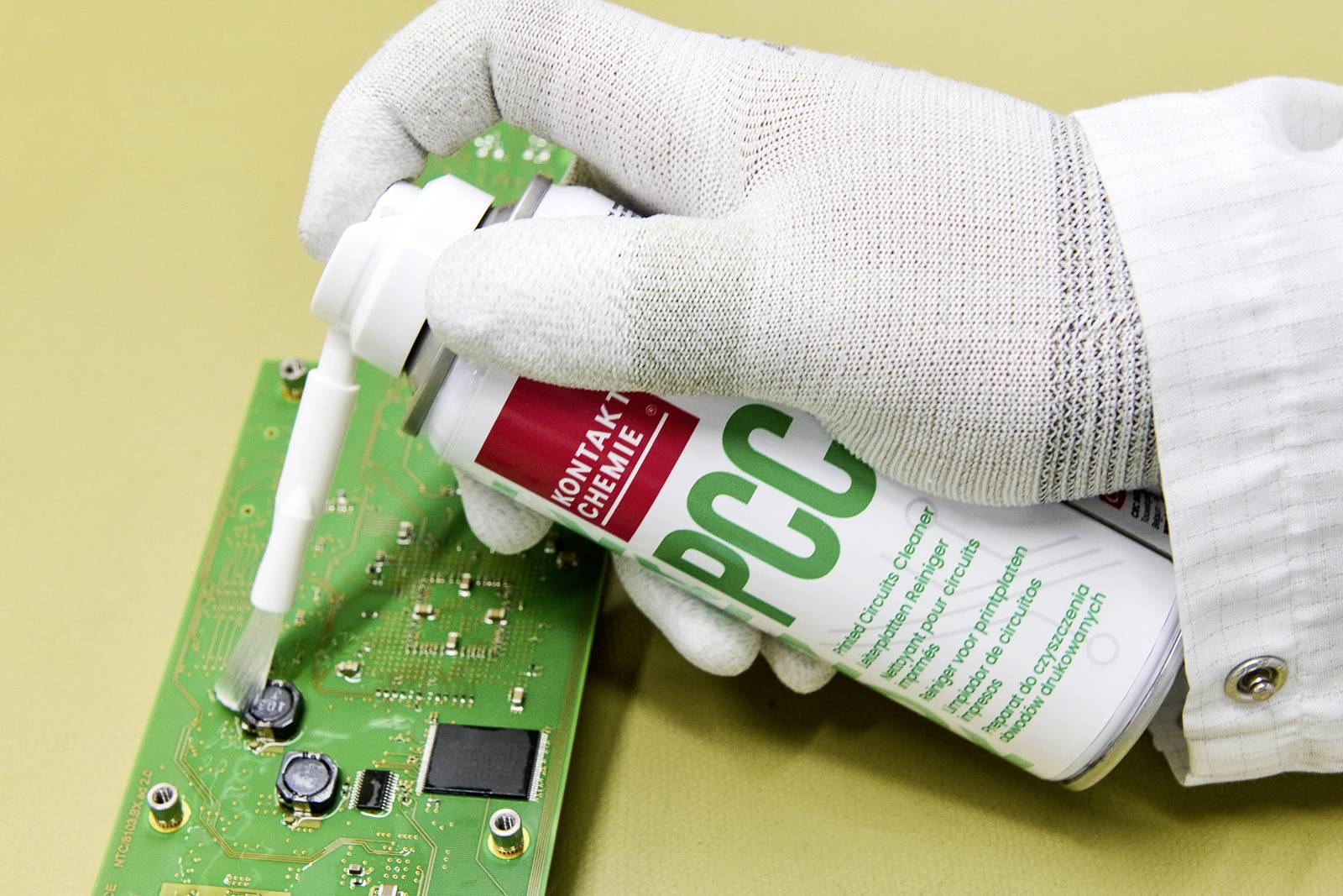 Una mano guantata spruzza un detergente su una scheda elettronica verde per pulire o sottoporre a manutenzione componenti elettronici.