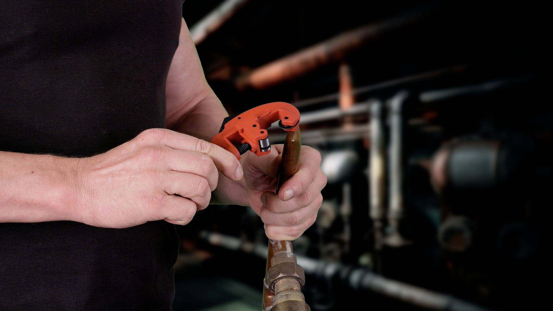 Una persona taglia un tubo di rame con un tagliatubi, sullo sfondo si intravedono sfocati impianti industriali.