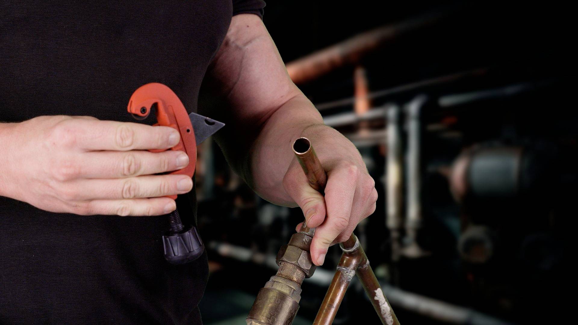 Una persona tiene un tubo di rame e un tagliatubi, sullo sfondo sfocato si vedono tubi e valvole. Rappresentazione di lavori di installazione.