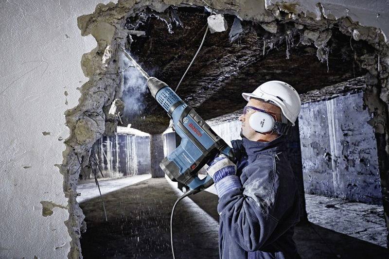 Un lavoratore edile con indumenti di sicurezza utilizza un utensile elettrico per forare un muro in calcestruzzo, creando un'apertura tra due stanze.
