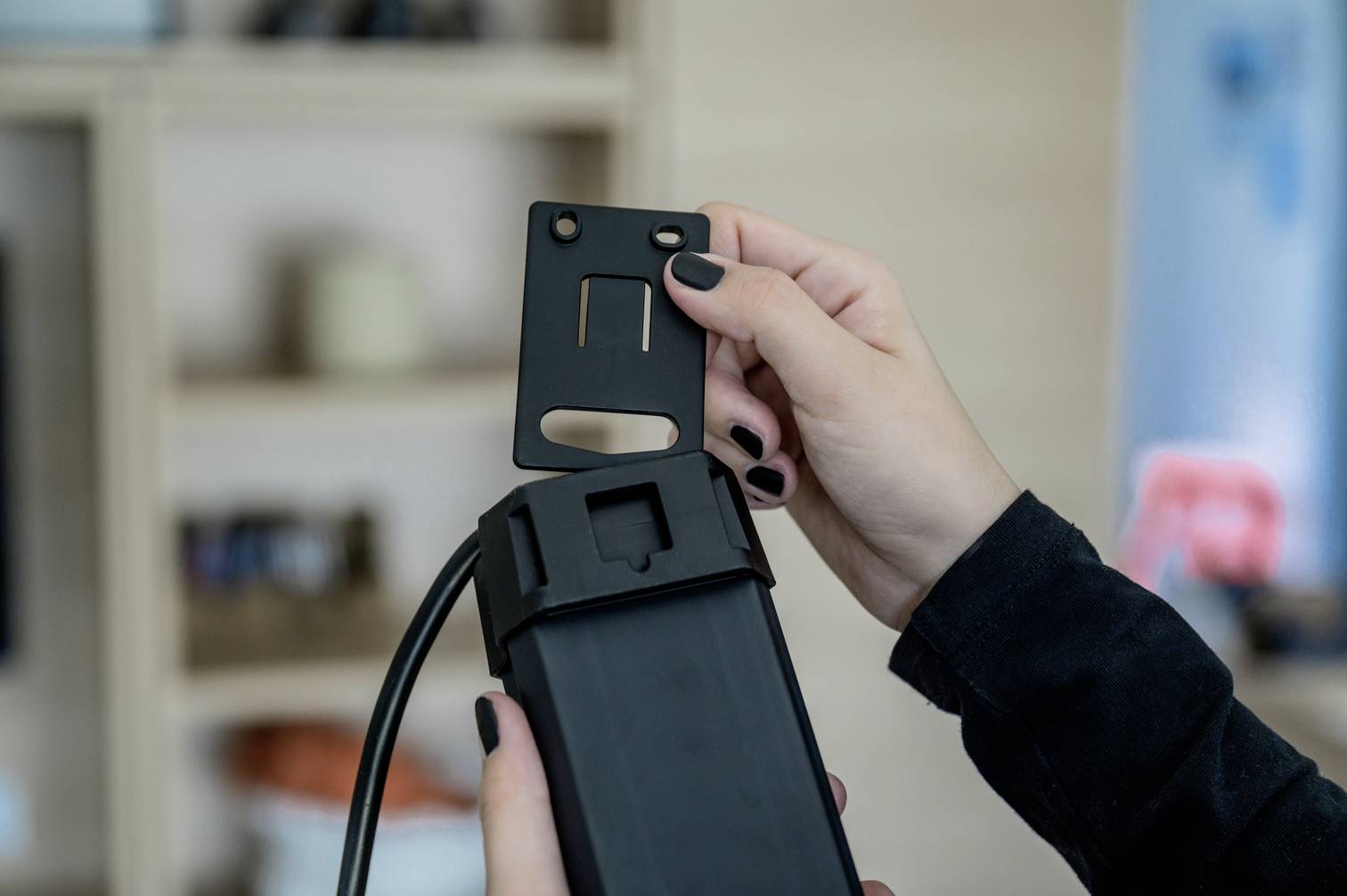 Una persona tiene un componente nero in plastica e un coperchio. Sullo sfondo sono visibili scaffali sfocati.