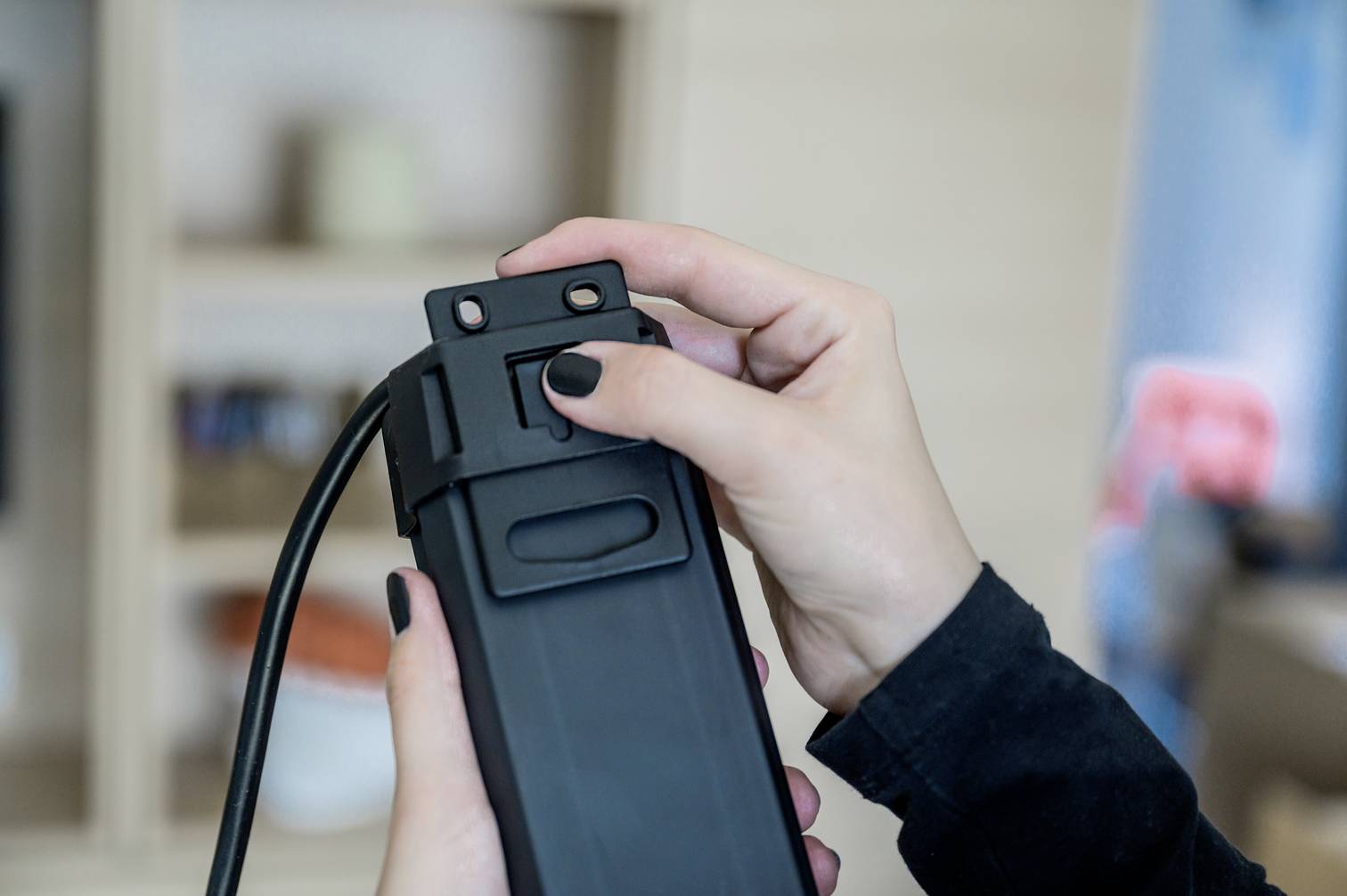 Una persona tiene e preme un interruttore su un dispositivo elettrico rettangolare. Sullo sfondo si intravedono mobili sfocati.