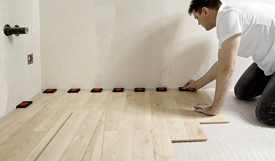 Una persona sta posando pannelli di legno per pavimento in una stanza, utilizzando distanziatori a parete per un allineamento preciso.