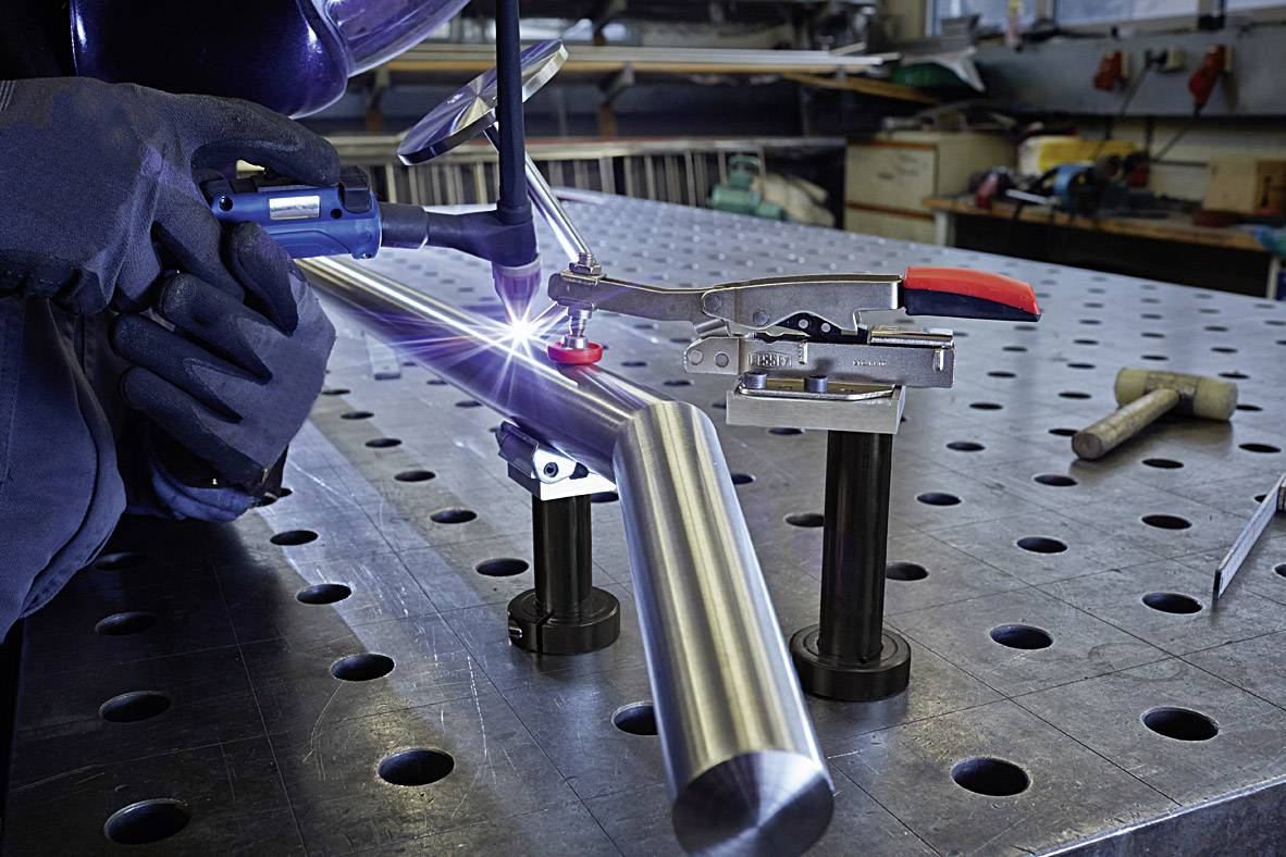 Una persona salda un tubo metallico su un tavolo da lavoro in officina, con indumenti protettivi e attrezzi sullo sfondo.