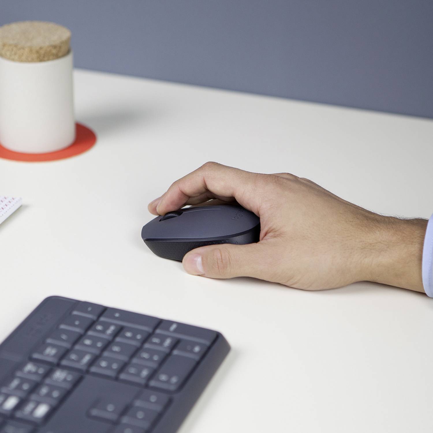 Una mano tiene un mouse per computer su un tavolo bianco accanto a una tastiera. Sullo sfondo, una tazza è poggiata su un sottobicchiere rosso.