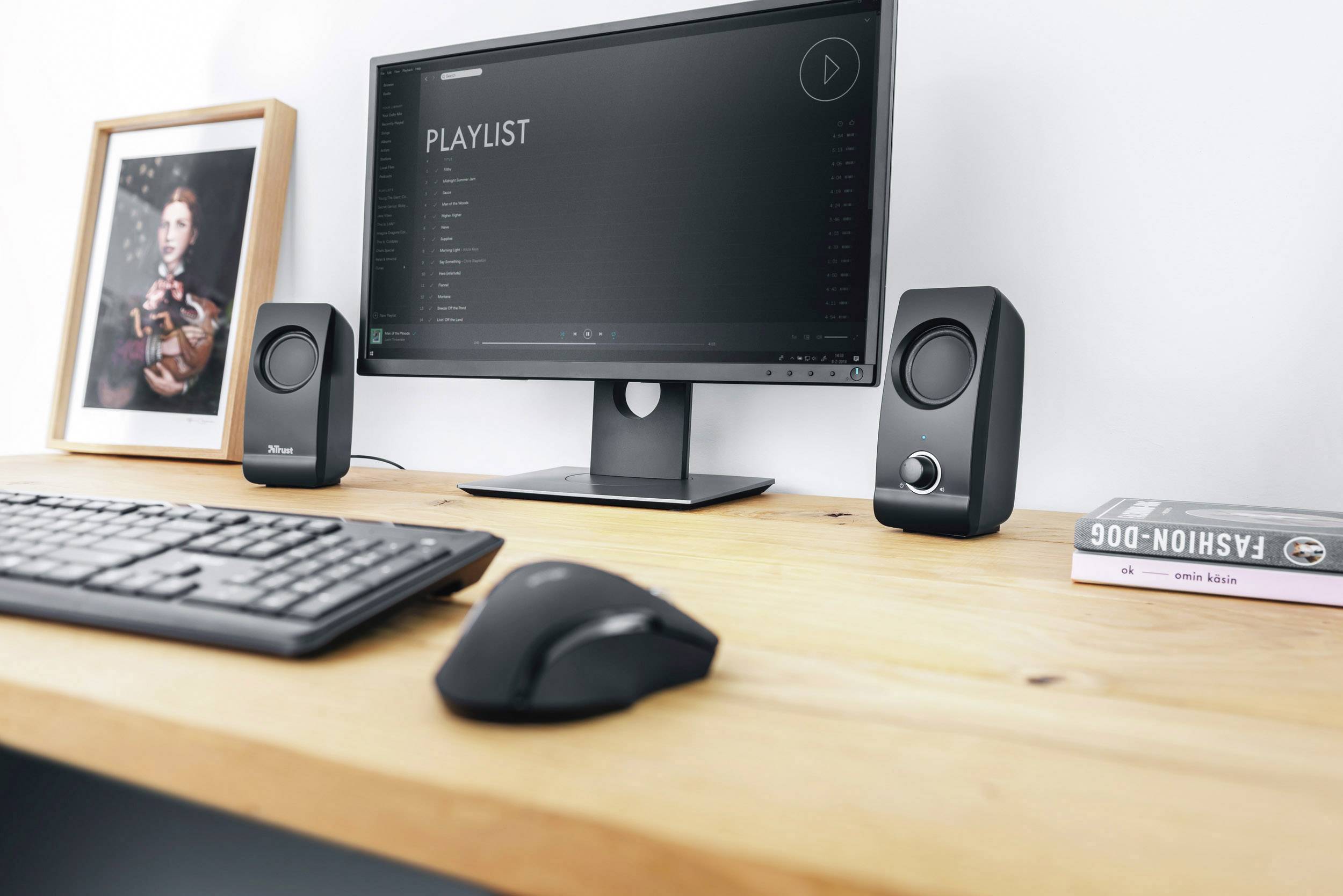 Un tavolo da lavoro con un monitor e altoparlanti mostra una playlist. Accanto alla tastiera c'è un mouse, a destra è posizionato un libro.