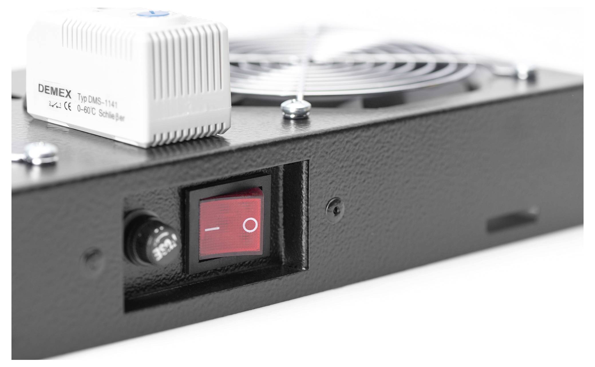 Un primo piano di un dispositivo elettronico nero con un interruttore rosso di accensione/spegnimento e un componente DEMEX bianco con una presa d'aria sullo sfondo.