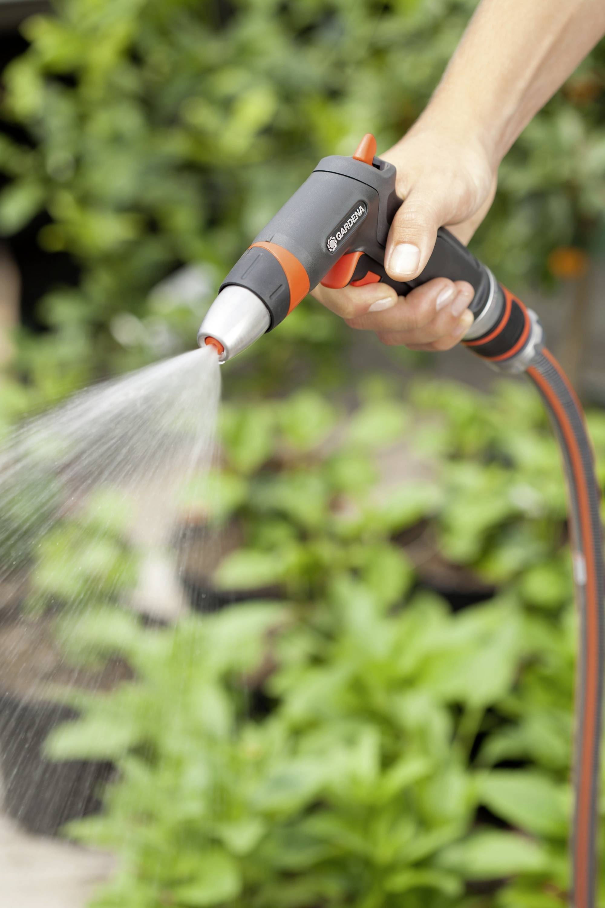 Una mano tiene un tubo da giardino con un ugello a spruzzo e irriga piante sullo sfondo.