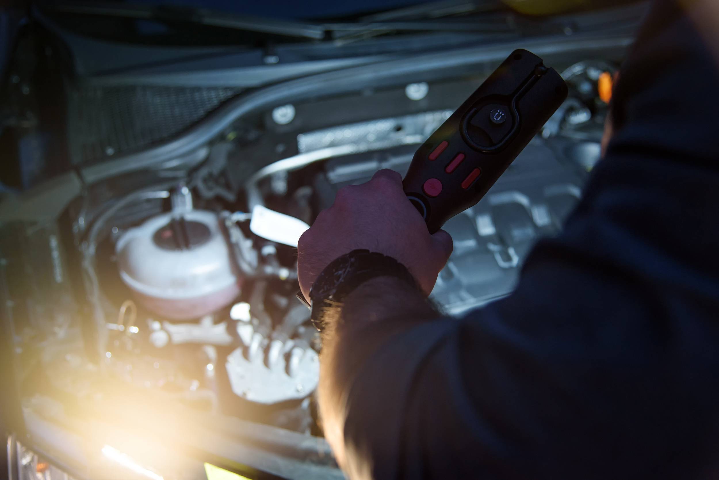 Una persona tiene un dispositivo diagnostico sopra il vano motore di un'auto aperto, per analizzare o riparare problemi tecnici.
